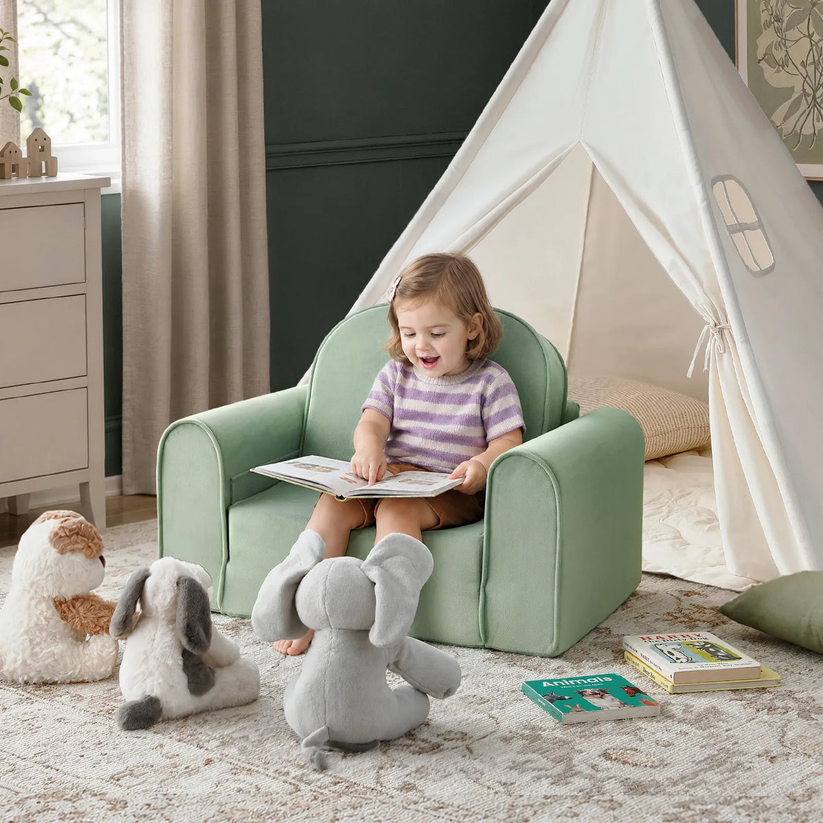 A young child sits on a Tiny Land® GentleSnug Kids Sofa Chair by Tiny Land, reading a book aloud to three stuffed animals in front of a white play tent; books are scattered on the floor nearby, creating a playful scene with cozy playroom furniture.|Light Green