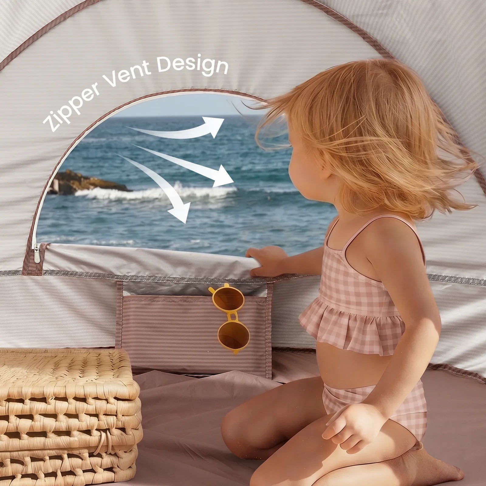A young child in a plaid swimsuit sits inside the Tiny Land® 4-in-1 Baby Beach Tent & Sand Toy Set, facing the ocean. The tent features a zippered vent for airflow, with sunglasses in its pocket and a woven basket nearby—perfect for beach play.|Pink
