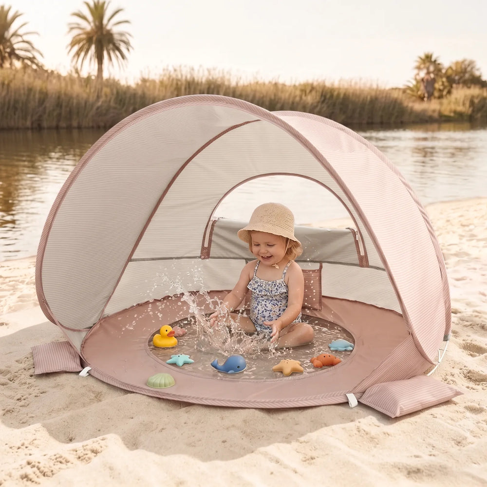 A young child in a sun hat enjoys sand and water play with floating toys in the Tiny Land® 4-in-1 Baby Beach Tent & Sand Toy Set by Tiny Land, set up on a sandy shore surrounded by palm trees and reeds.