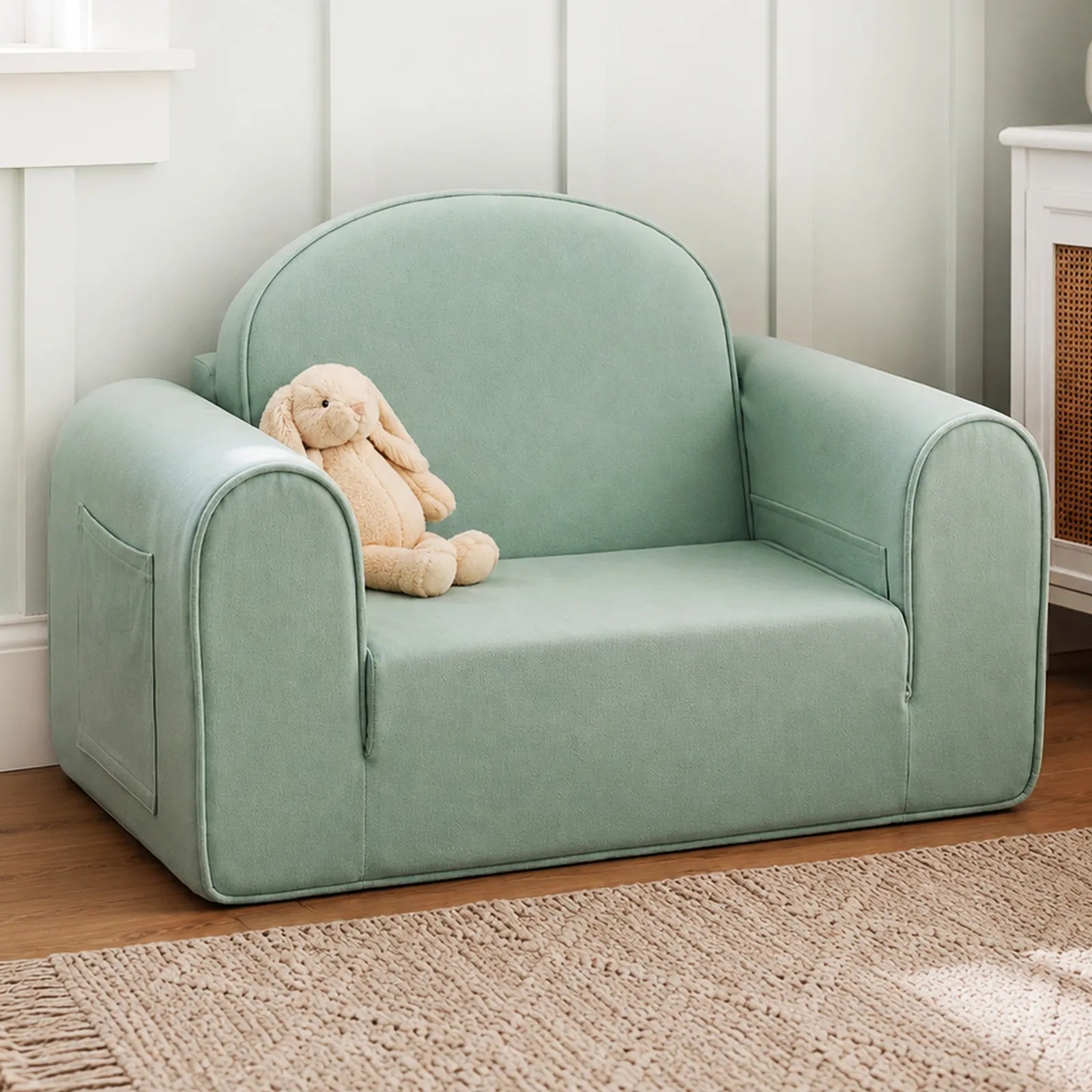 The Tiny Land® GentleSnug Kids Sofa Chair by Tiny Land, in light green with wide arms and a side pocket, sits on a wooden floor; a beige stuffed bunny rests on the left side of the toddler chair.