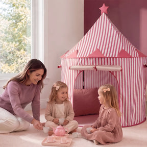 A woman and two young girls have a tea party on the floor beside a Tiny Land® Kids Tent – Indoor Tent for Toddlers, featuring pink stripes and a star on top, in a softly lit room with a large window.|Pink
