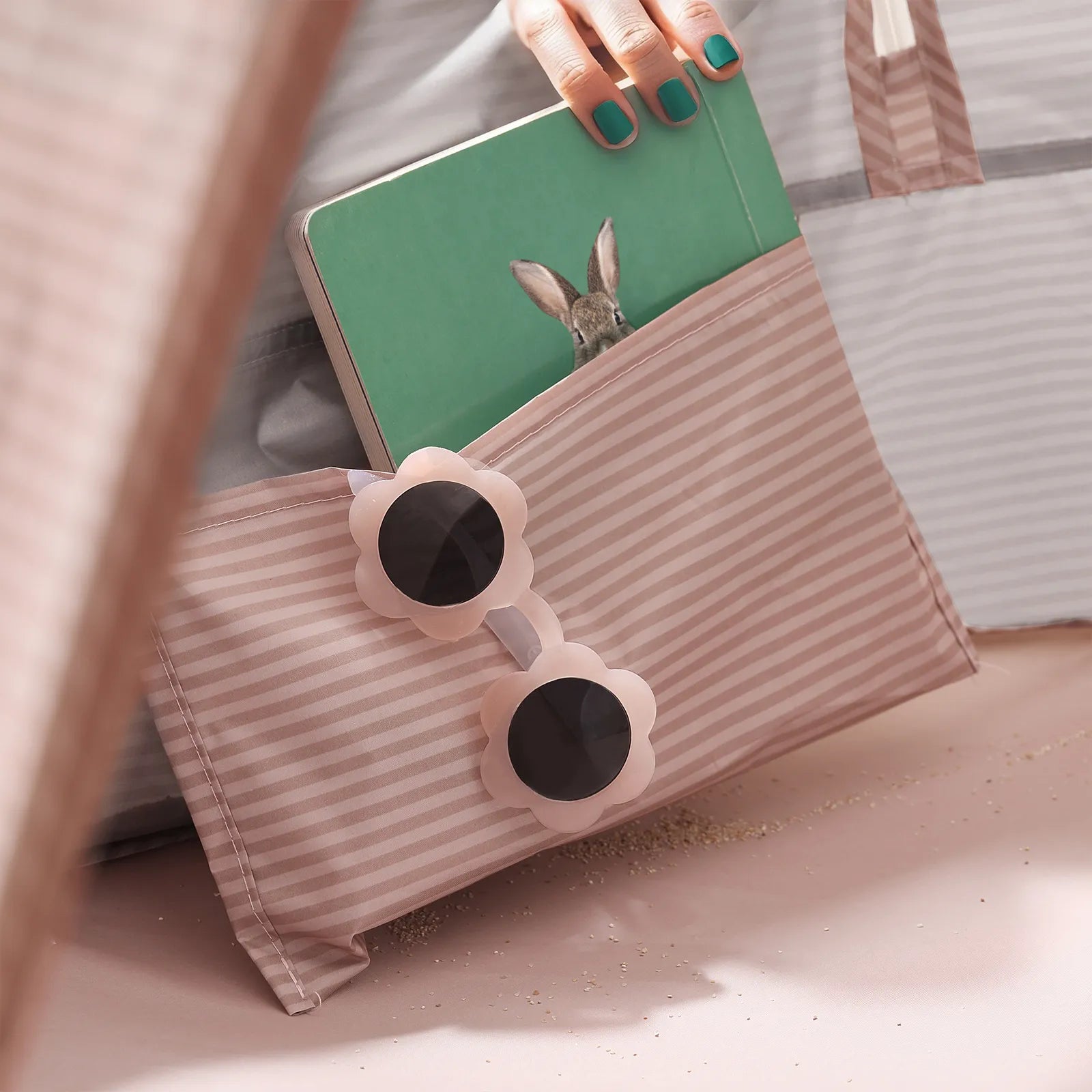 A hand with teal nail polish holds a green book with a rabbit cover, partly tucked into a pink-striped bag beside flower sunglasses—perfect with the Tiny Land® 4-in-1 Baby Beach Tent & Sand Toy Set for toddler beach fun.|Pink