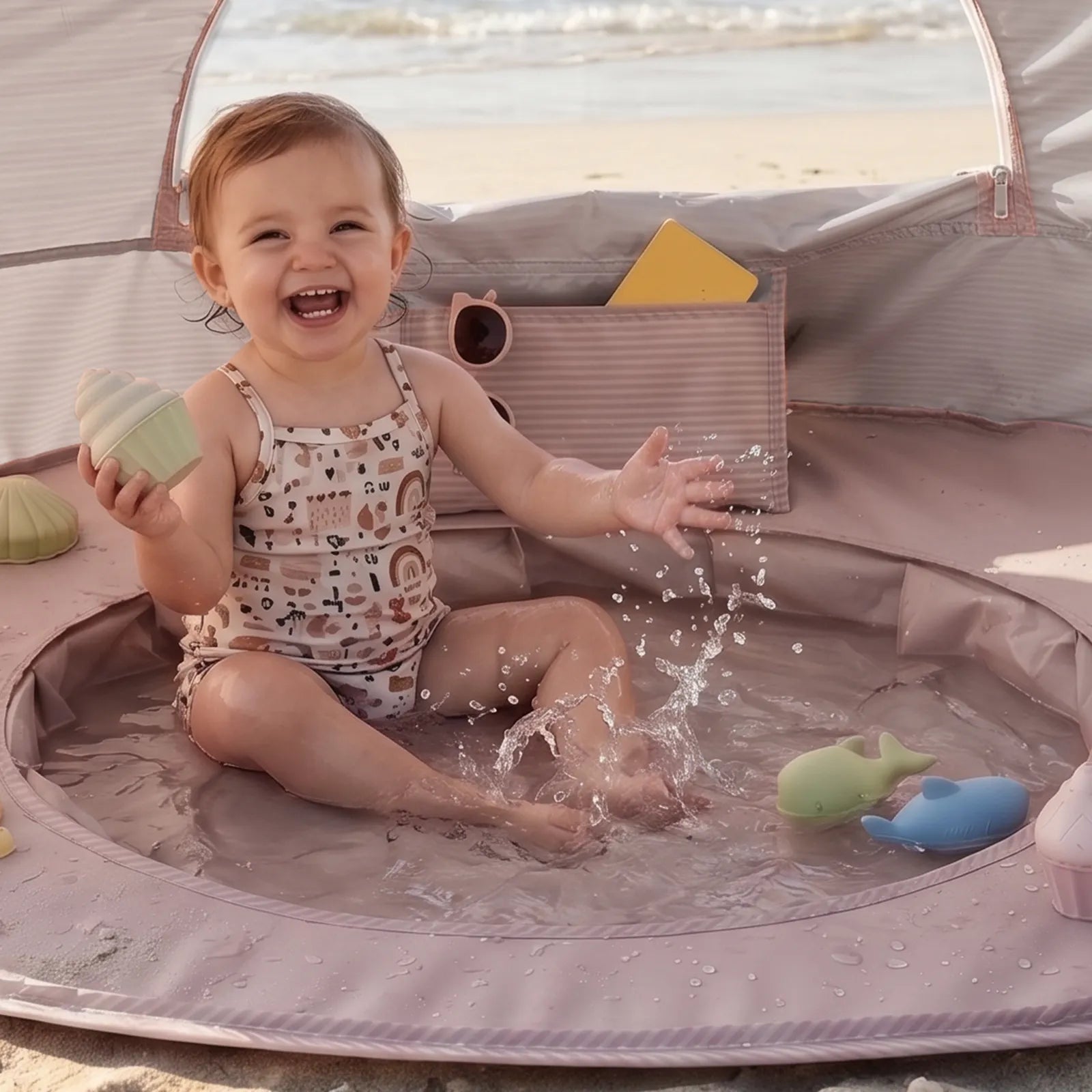 A happy toddler splashes in a Tiny Land® 4-in-1 Baby Beach Tent & Sand Toy Set, surrounded by floating toy animals and shapes, with a handy pocket for toys—perfect for endless sand and water fun on the beach.|Pink