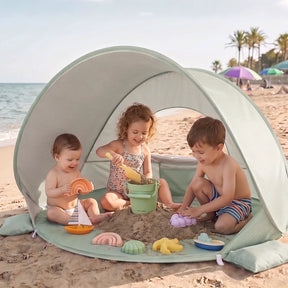 Three young children play with buckets and beach toys under the Tiny Land® 4-in-1 Baby Beach Tent & Sand Toy Set. The light green tent sits on the sand, with umbrellas and palm trees in the background, ideal for sand and water play.|Green