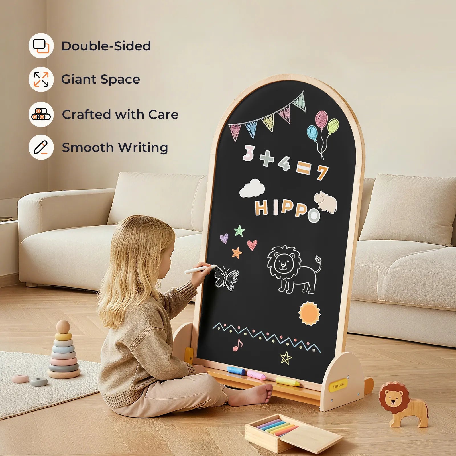 A young child draws with chalk on the Tiny Land® Double-Sided Arched Magnet Chalkboard, featuring math, balloons, and animal doodles. Wooden Tiny Land lion toy and stacked rings rest nearby on the wooden floor.