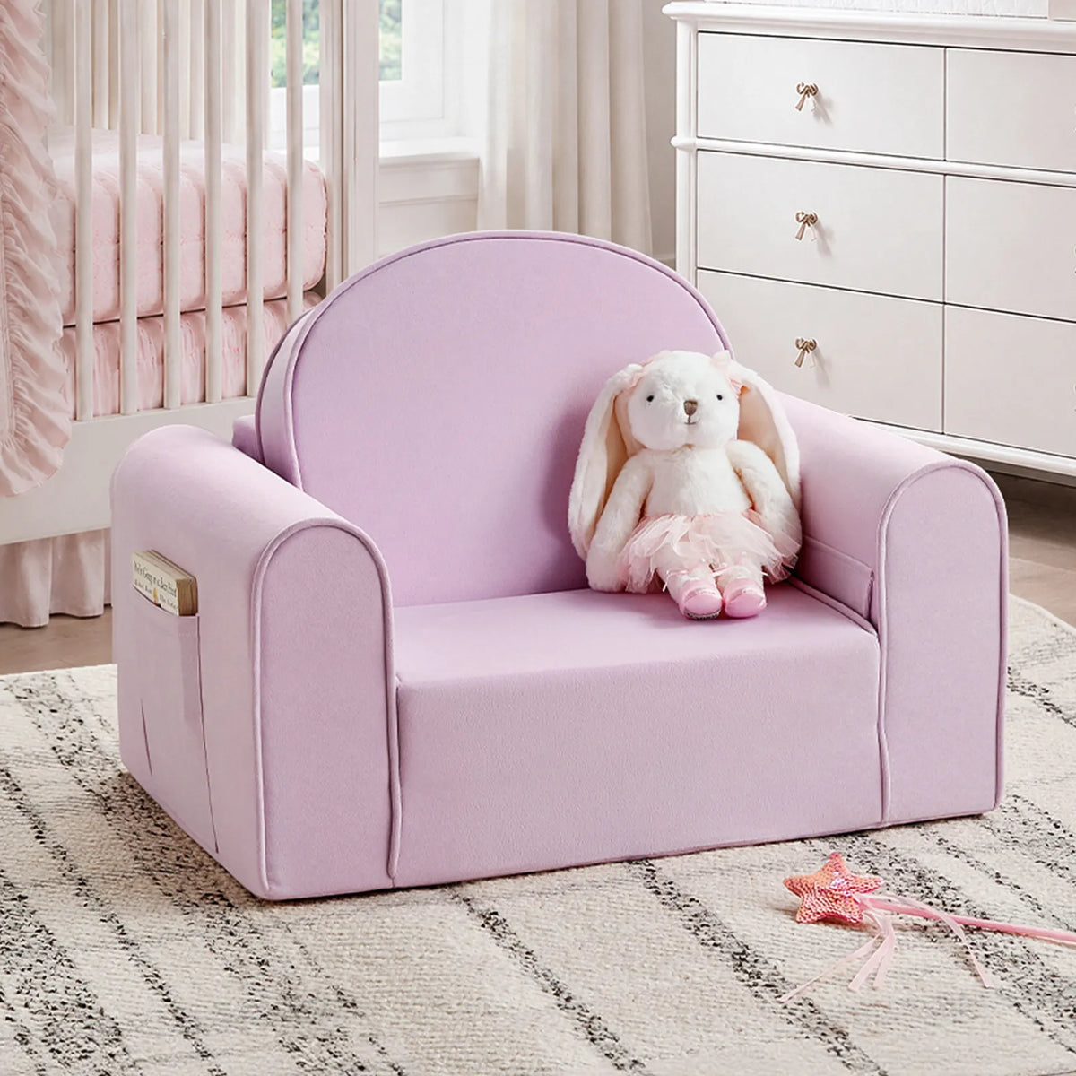 The Tiny Land® GentleSnug Kids Sofa Chair—a pink toddler sofa by Tiny Land—is shown with a cuddly white rabbit in a matching pink dress seated on it, set atop a patterned rug in a nursery complete with crib and dresser. It’s the perfect playful addition to your little one’s playroom or bedroom furniture collection.|Light Purple