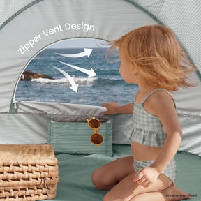 A young child in a checkered swimsuit sits inside the Tiny Land® 4-in-1 Baby Beach Tent & Sand Toy Set, peering out at the ocean. Sunglasses hang on the Tiny Land tent, with a woven basket nearby for sand and water play.|Green