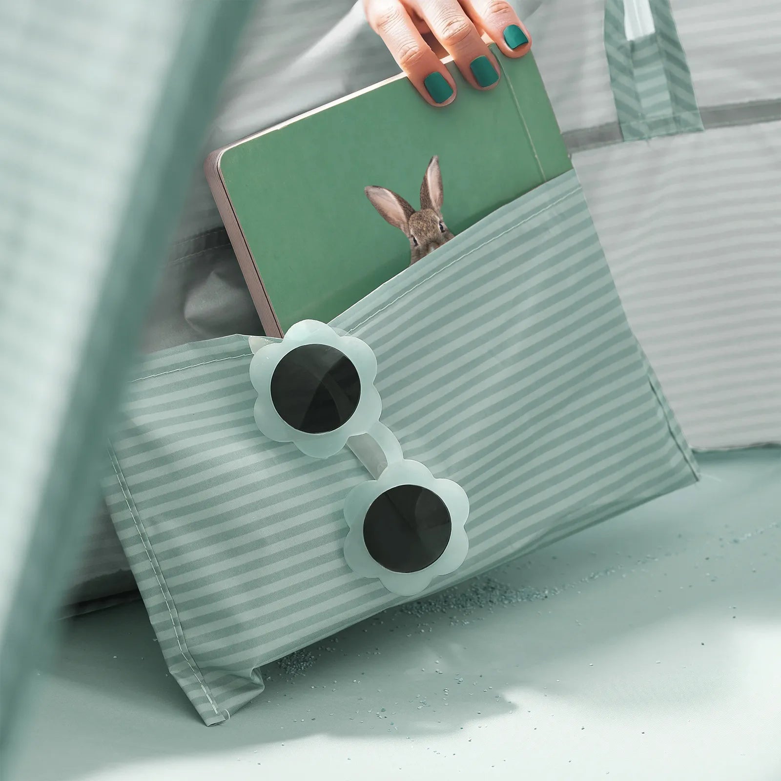 A hand with teal nail polish holds a green tablet with a rabbit image, partially tucked in a striped mint pouch. Flower-shaped sunglasses rest nearby—perfect accessories for the Tiny Land® 4-in-1 Baby Beach Tent & Sand Toy Set by Tiny Land.|Green
