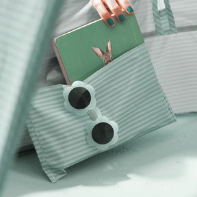 A hand with teal nail polish holds a green tablet with a rabbit image, partially tucked in a striped mint pouch. Flower-shaped sunglasses rest nearby—perfect accessories for the Tiny Land® 4-in-1 Baby Beach Tent & Sand Toy Set by Tiny Land.|Green