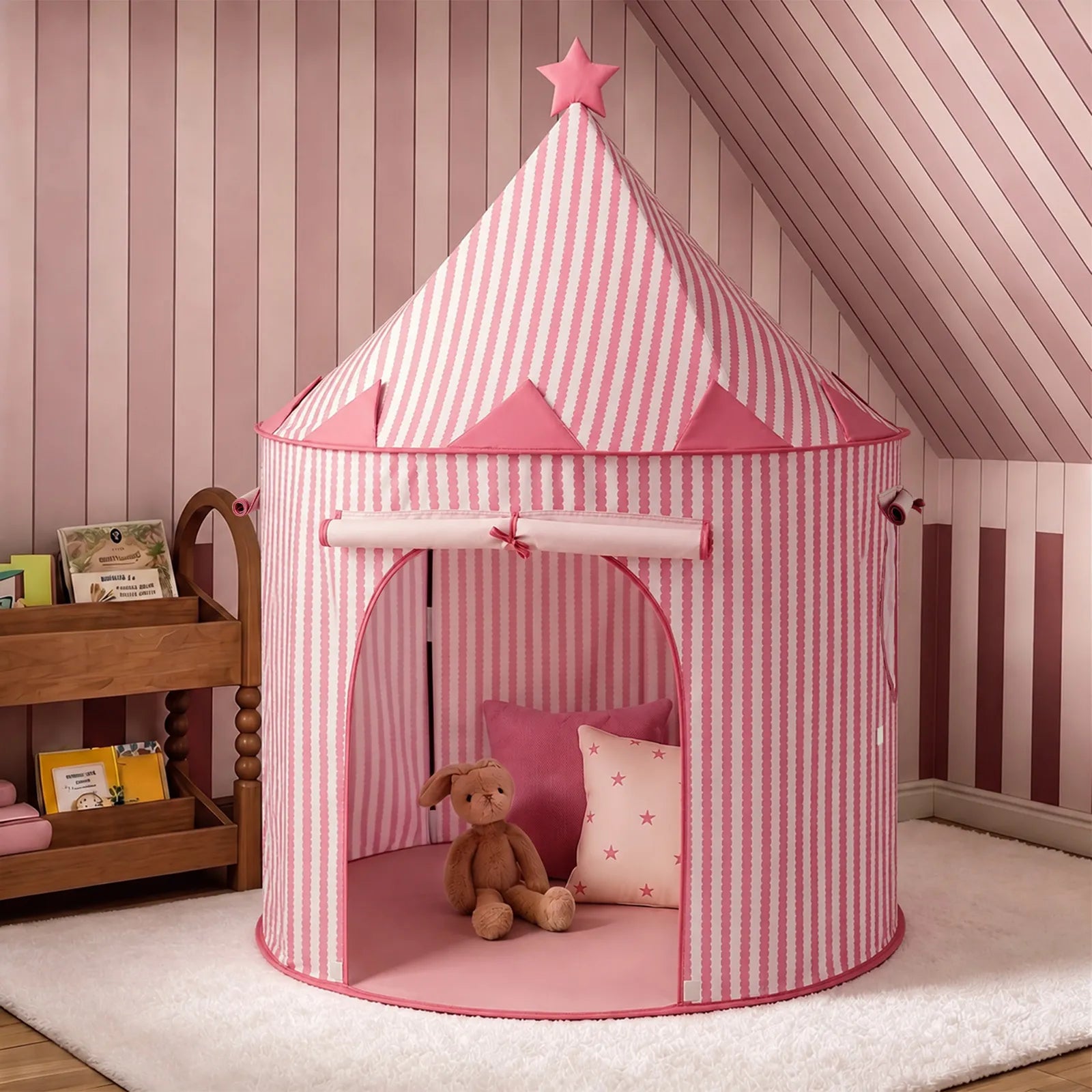 A Tiny Land® Kids Tent – Indoor Tent for Toddlers in pink and white stripes, topped with a star, stands in a pink room. Inside the tent are a brown stuffed bunny and a pink pillow decorated with stars. A wooden shelf holding books is placed nearby.|Pink