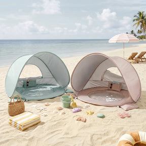Two Tiny Land® 4-in-1 Baby Beach Tent & Sand Toy Sets, one mint green and one beige, with built-in pools for sand and water play, sit on the beach surrounded by toys, towels, and chairs; palm trees and the ocean are in view.