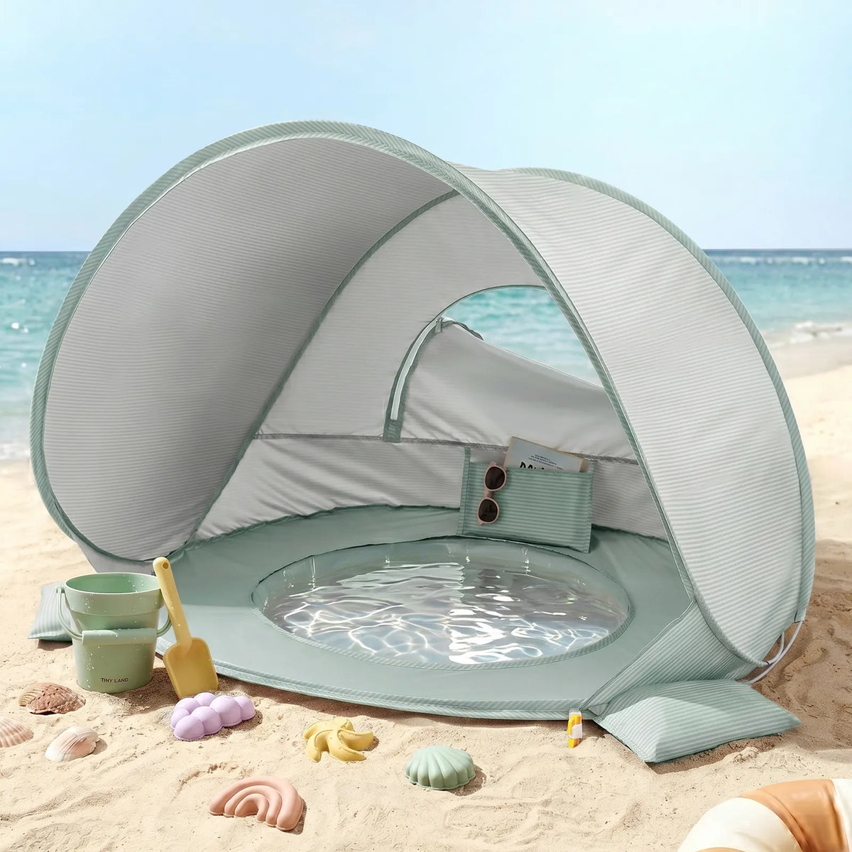 The Tiny Land® 4-in-1 Baby Beach Tent & Sand Toy Set by Tiny Land is set up on a sandy beach near the ocean, featuring a shaded canopy, built-in mini pool, beach toys, and seashell molds for endless sand and water play with the sea in the background.|Green