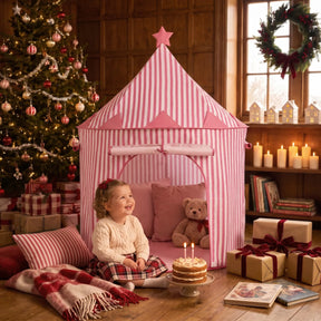 A smiling toddler sits in front of the Tiny Land® Kids Tent – Indoor Tent for Toddlers by Tiny Land, with a teddy bear nearby, surrounded by wrapped gifts, a cake with candles, a Christmas tree, and festive decorations in a cozy room. The cheerful Tiny Land tent adds an extra touch of magic to the holiday celebration.|Pink