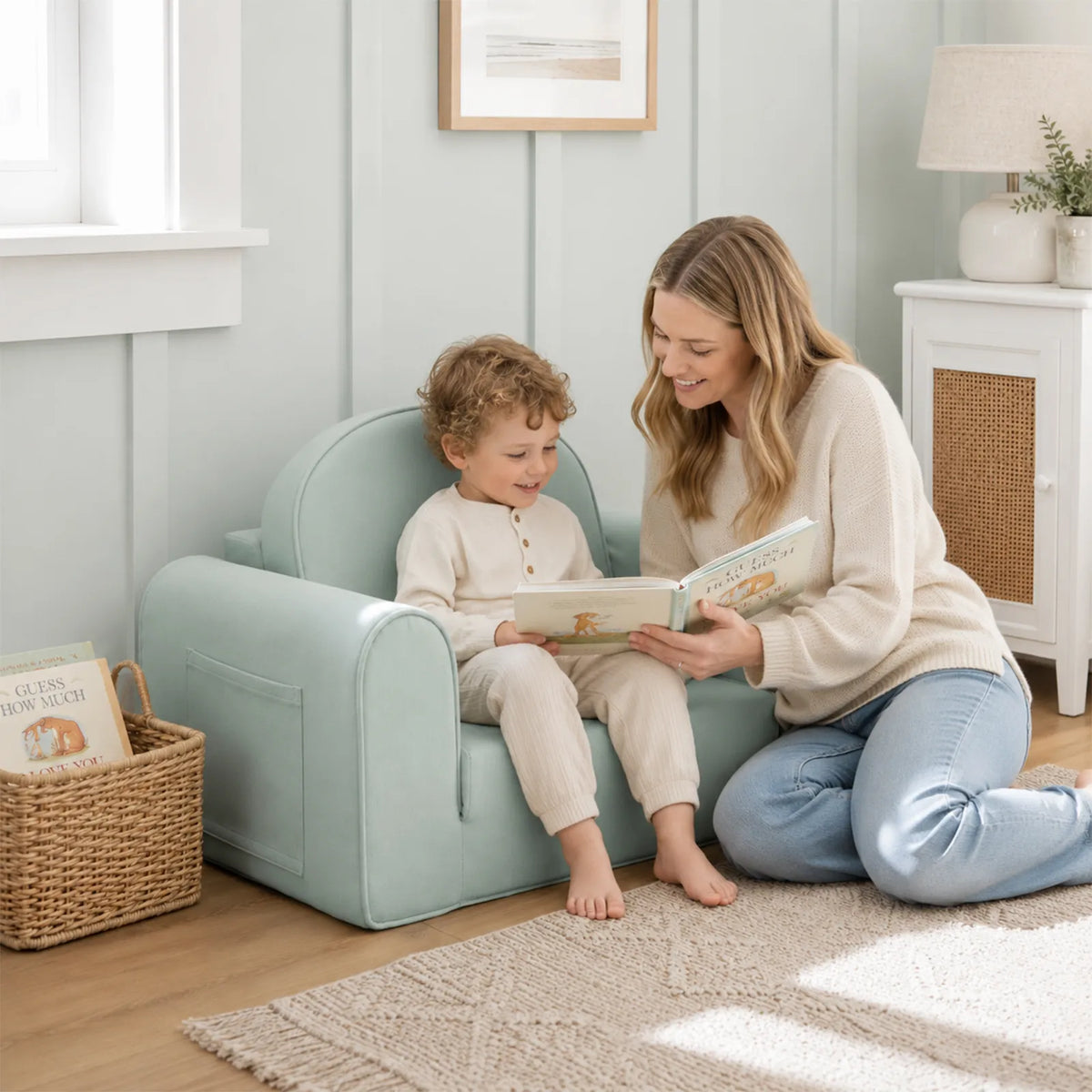 A woman kneels on a rug and reads to a young child sitting on a Tiny Land® GentleSnug Kids Sofa Chair in a sunlit room with books, soft decor, and a basket.