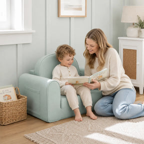 A woman kneels on a rug and reads to a young child sitting on a Tiny Land® GentleSnug Kids Sofa Chair in a sunlit room with books, soft decor, and a basket.