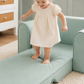 A young child in a cream-colored dress balances on the Tiny Land® GentleSnug Kids Sofa Chair, a light green fold-out cushion seat by Tiny Land, in a cozy room with a rug and dresser.