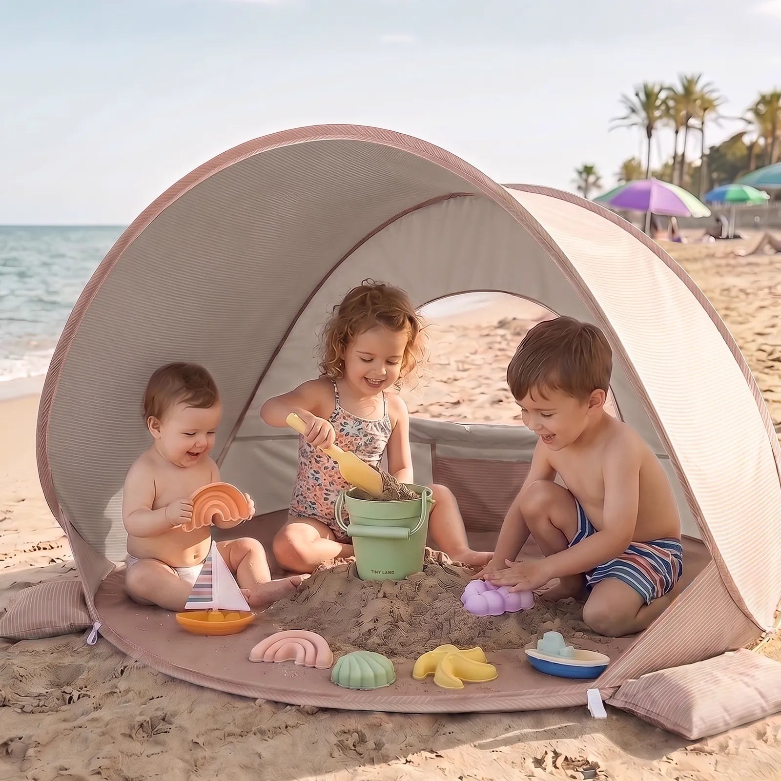 Three kids play with colorful sand and water toys under the Tiny Land® 4-in-1 Baby Beach Tent & Sand Toy Set by Tiny Land on a sunny beach, with the ocean and palm trees in the background.|Pink