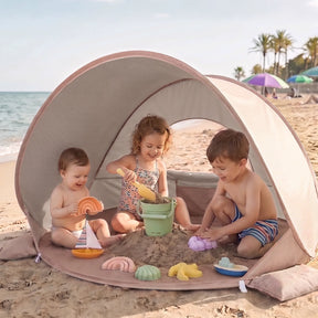 Three kids play with colorful sand and water toys under the Tiny Land® 4-in-1 Baby Beach Tent & Sand Toy Set by Tiny Land on a sunny beach, with the ocean and palm trees in the background.|Pink