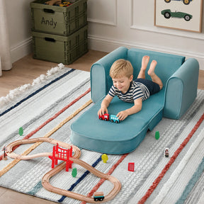A young boy relaxes on the Tiny Land® GentleSnug Kids Sofa Chair by Tiny Land, its pull-out footrest extended, as he plays with toy trains on a wooden track set atop a striped rug in a cozy room adorned with car-themed art.|Blue