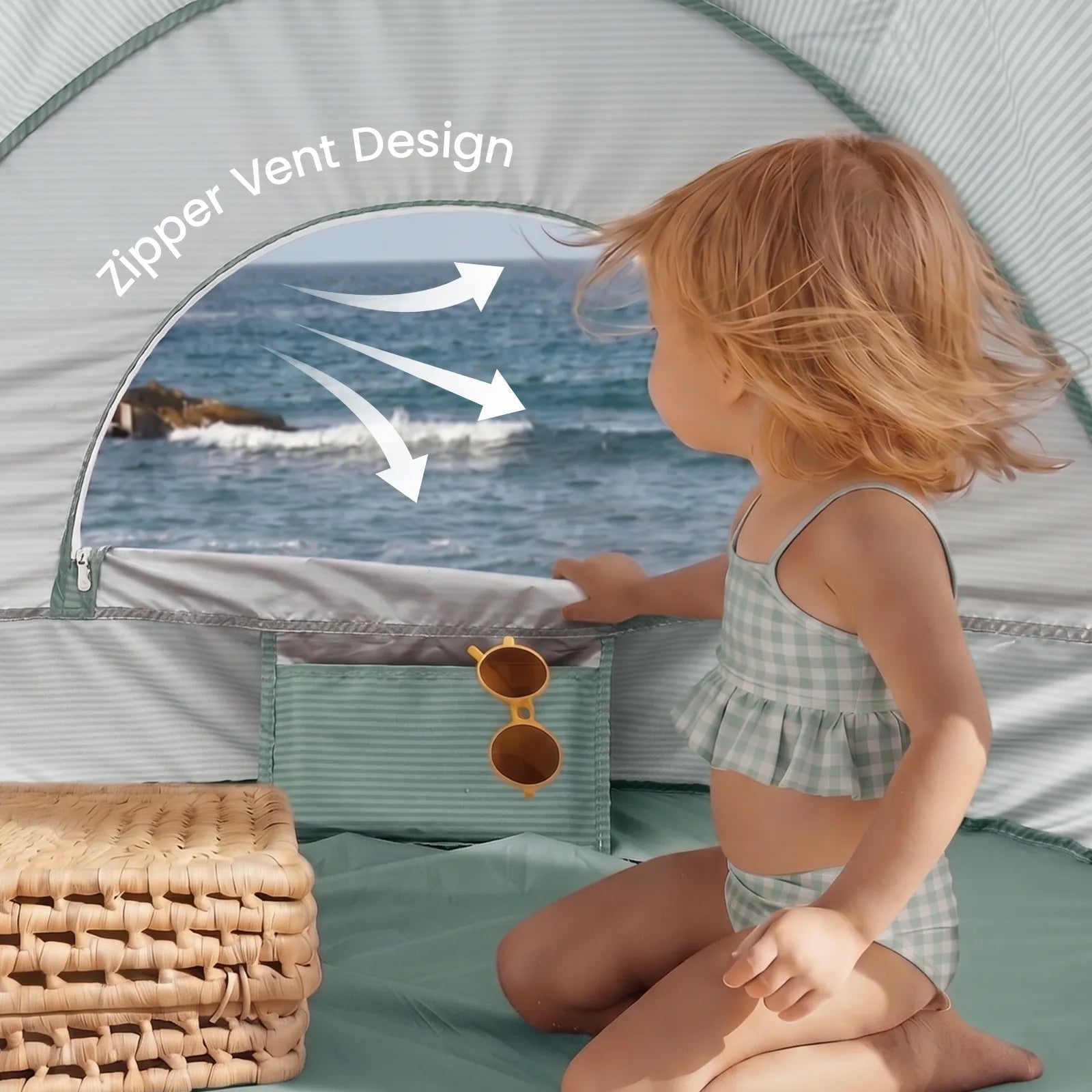 A young child in a checkered swimsuit sits inside the Tiny Land® 4-in-1 Baby Beach Tent & Sand Toy Set on the sand, peering out a zipper vent. Sunglasses and a wicker basket nearby await beach play with the Tiny Land set.|Green