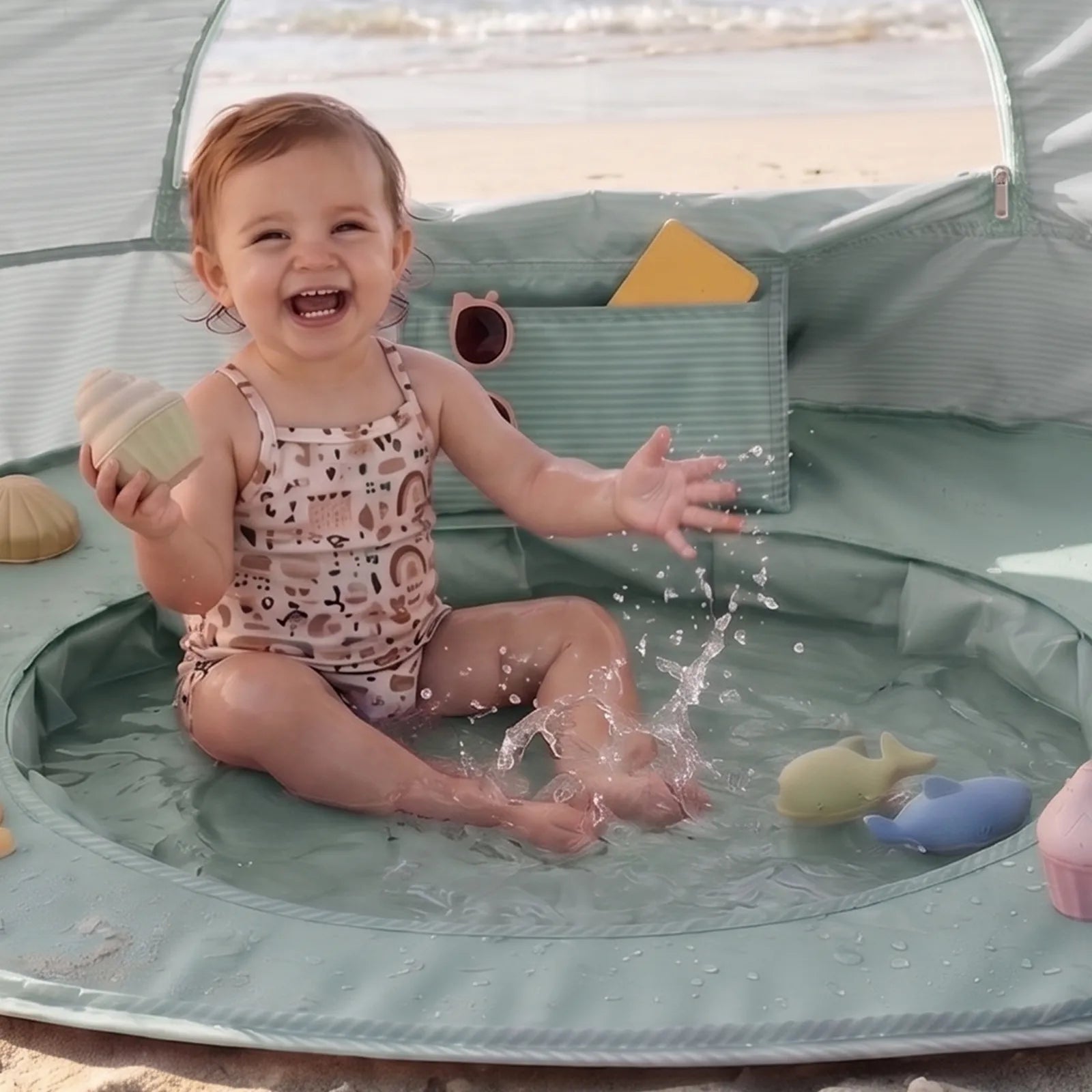 A happy toddler plays in the Tiny Land® 4-in-1 Baby Beach Tent & Sand Toy Set, splashing water and enjoying colorful sea creature toys on the sand, with the ocean in the background—ideal for fun sand and water play.|Green