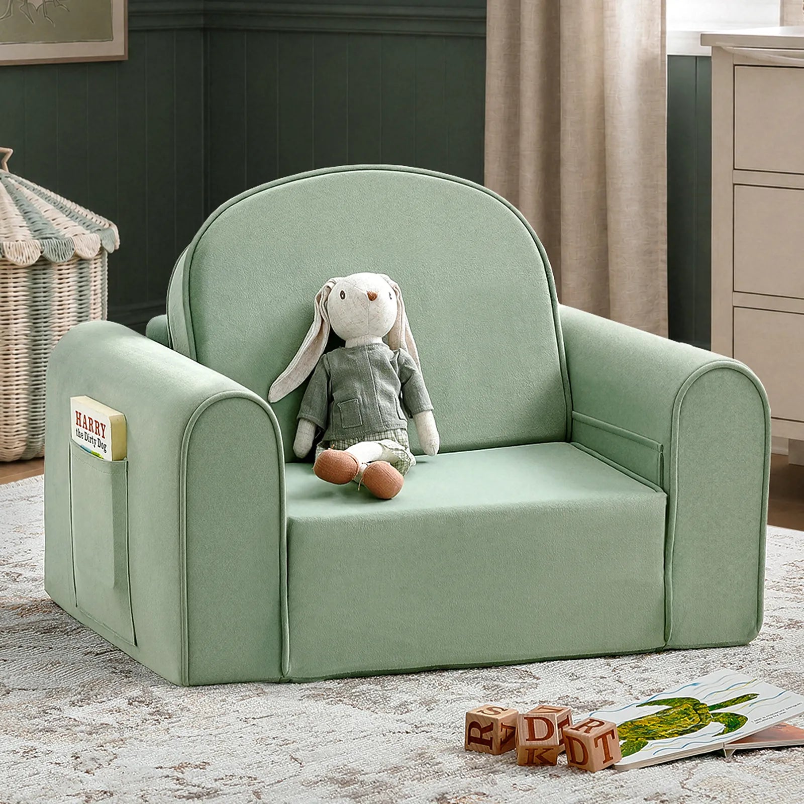 The Tiny Land® GentleSnug Kids Sofa Chair, a small green toddler sofa, sits invitingly with a stuffed bunny toy perched on it. This cozy piece of playroom furniture from Tiny Land features side pockets—one holding a book—while wooden alphabet blocks and another book are scattered on the nearby floor.|Light Green