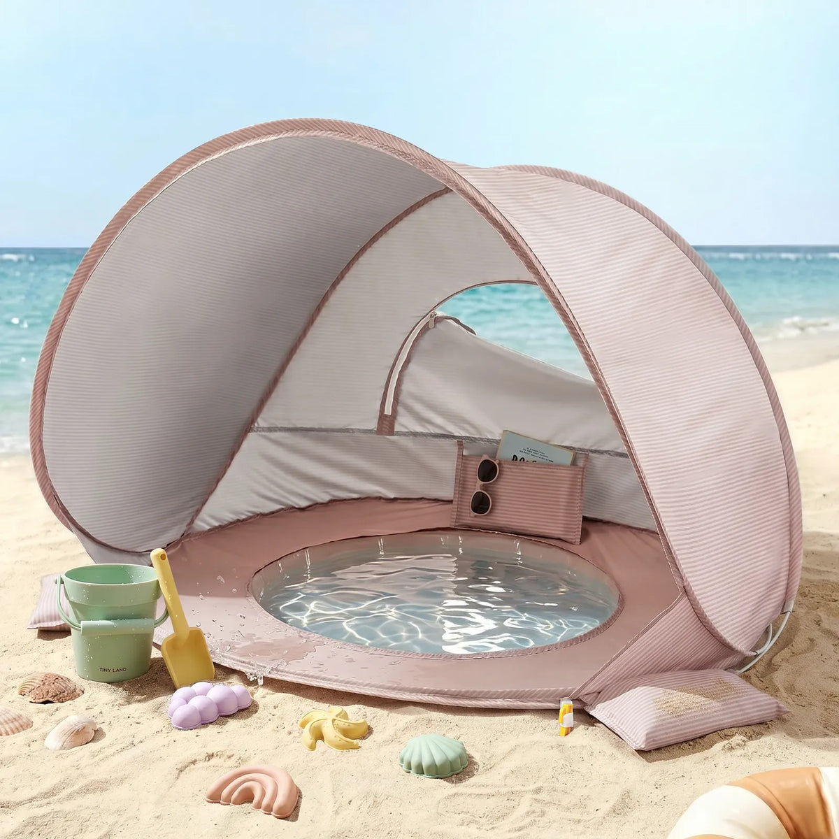 The Tiny Land® 4-in-1 Baby Beach Tent & Sand Toy Set by Tiny Land is set up on the beach near the sea, offering a built-in shallow pool and sand toys for fun sand and water play under a clear sky.|Pink