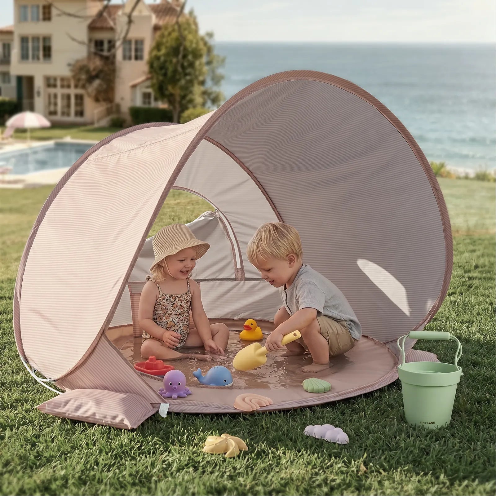 Two young children play with the Tiny Land® 4-in-1 Baby Beach Tent & Sand Toy Set on a lawn by the ocean. The shaded tent, from Tiny Land, shelters them as they enjoy sand and water play, with extra toys and a bucket outside by a house and pool.|Pink