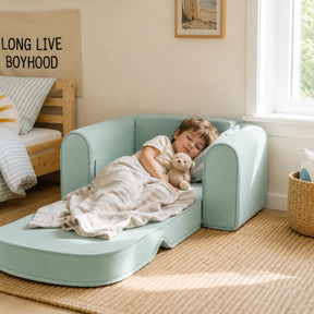 A young boy sleeps on a Tiny Land® GentleSnug Kids Sofa Chair, cuddling a teddy bear under a striped blanket in a cozy, sunlit room with a woven rug and a Long Live Boyhood pillow.
