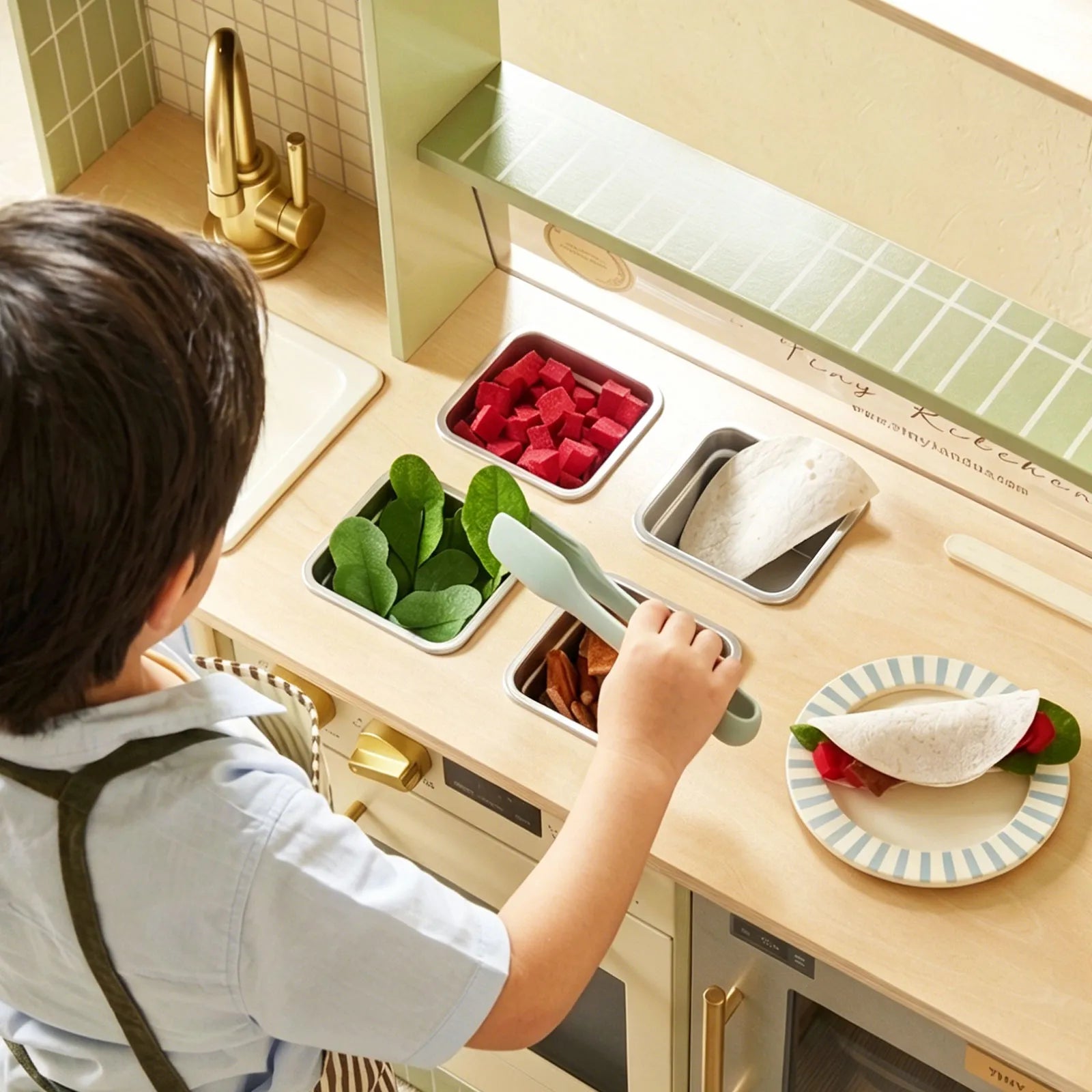 A child wearing an apron enjoys pretend play with the Tiny Land® BistroSwitch Play Kitchen, placing toy spinach and bread onto a plate near the sink and gold faucet—a delightful kids’ kitchen set by Tiny Land.|Green