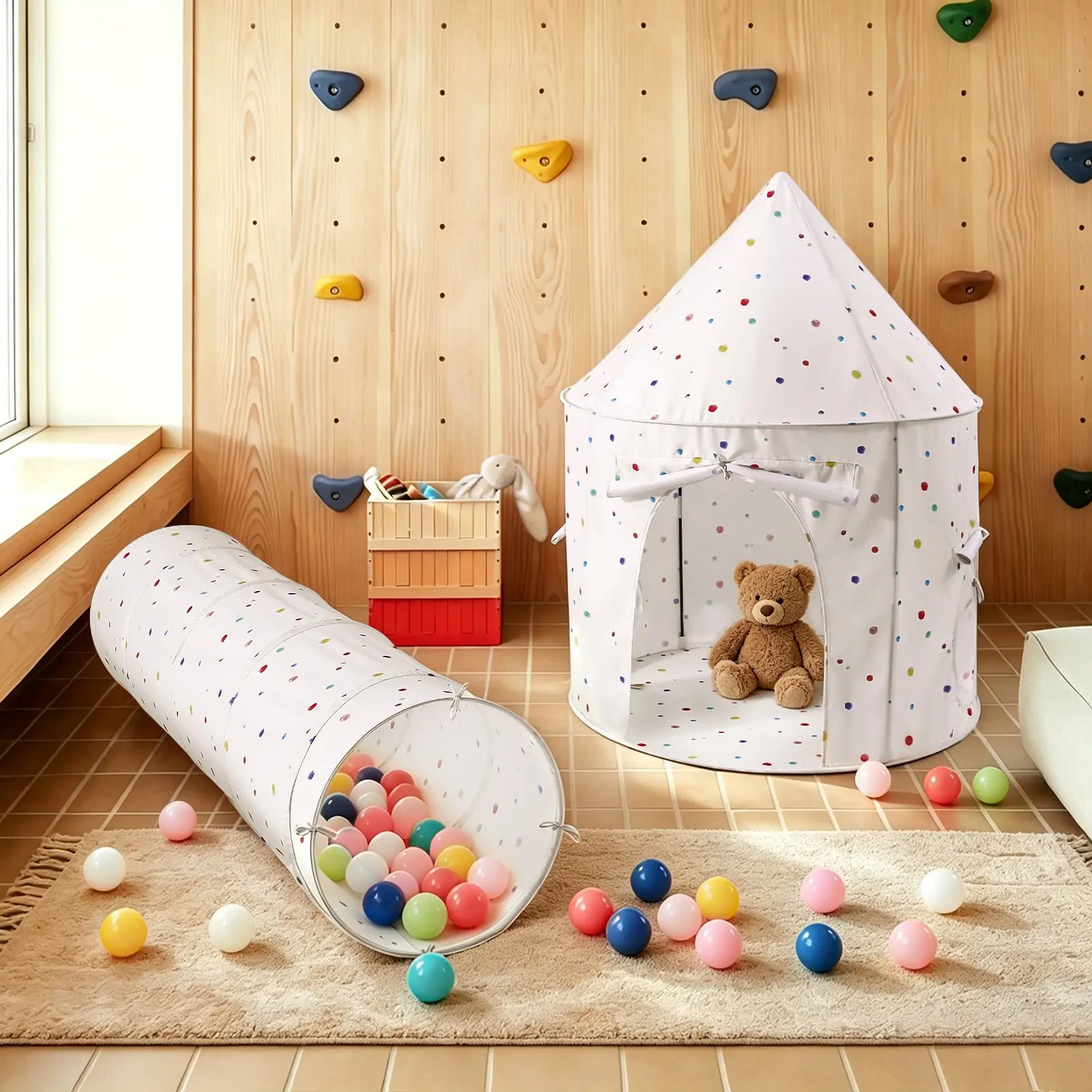 A cozy playroom features the Tiny Land® Adventure Play Tunnel - Colorful Polka Dots by Tiny Land filled with balls, a teddy bear inside, and a wooden wall in the background with climbing holds and toy storage.
