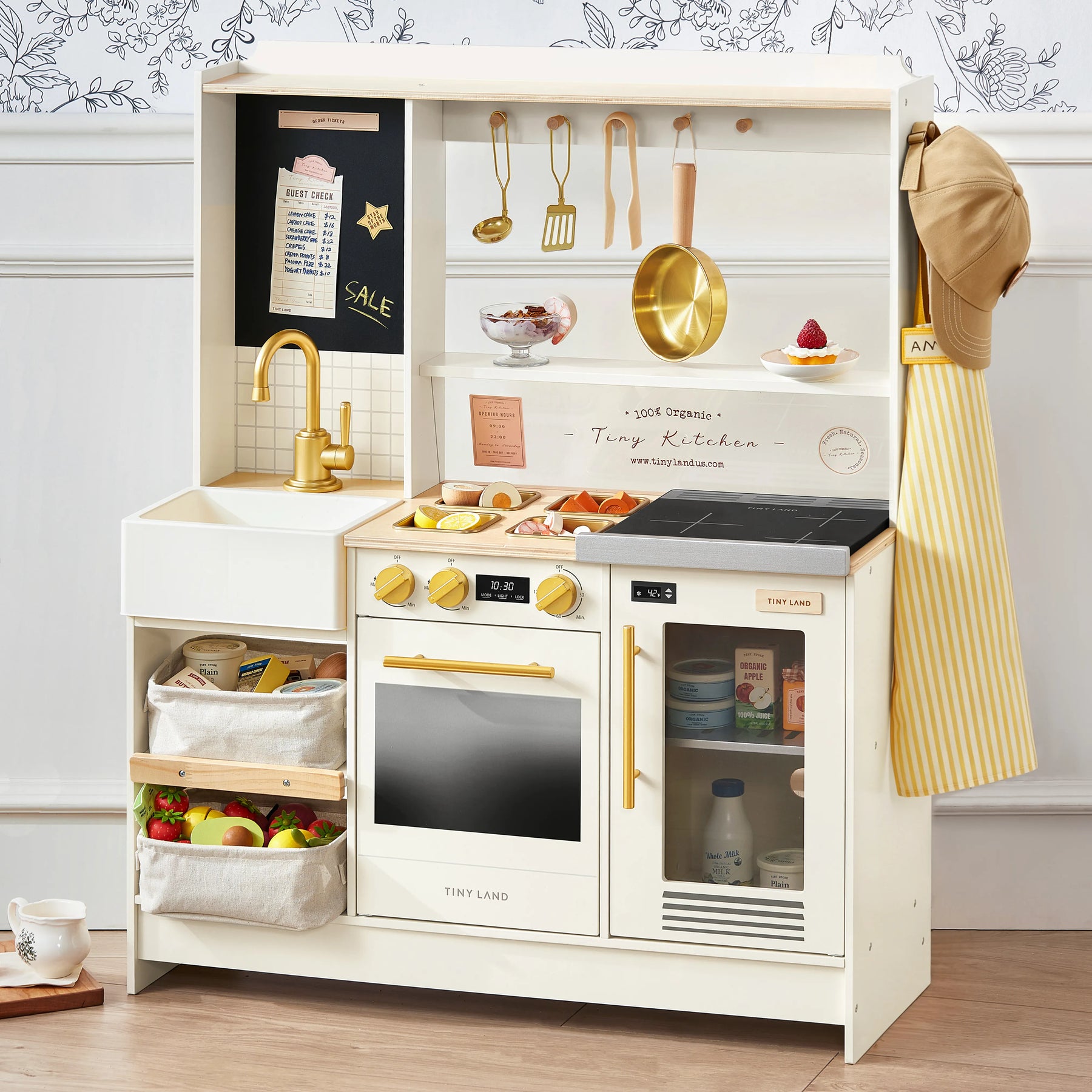 The Tiny Land® BistroSwitch Play Kitchen by Tiny Land features a sink, stove, oven, shelves, utensils and accessories above, play food like eggs and veggies on display, an apron and hat on the side, plus baskets with pretend fruits and vegetables.|White