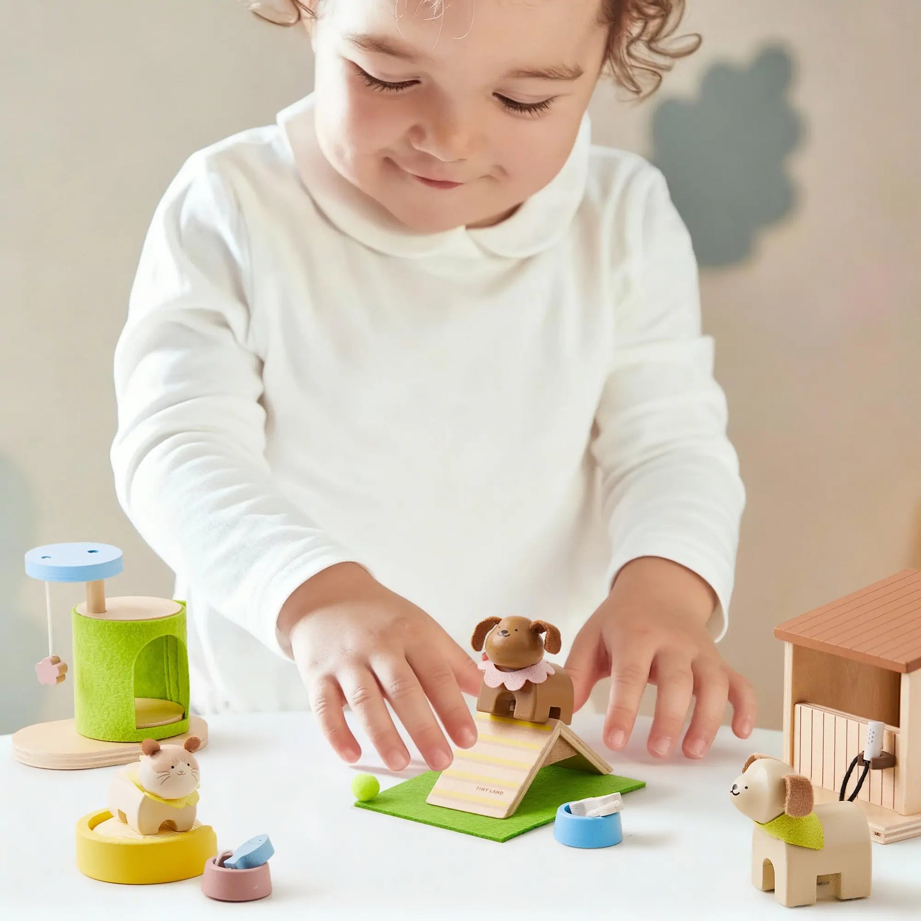 A young child in a white shirt smiles as they play with pretend pet toys and wooden animal figurines from the Test Early Bird Eggs-Tiny Land® Happy Urban Paws set, creating a playful scene on the table.