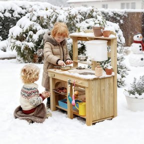 Two children play outside with the Tiny Land® Mud Kitchen - DuoPlay Outdoor Play Kitchen for Kids. One pretends to cook while the other watches, surrounded by snow-covered trees and a snowman in a wintry backyard. Brand: Tiny Land.