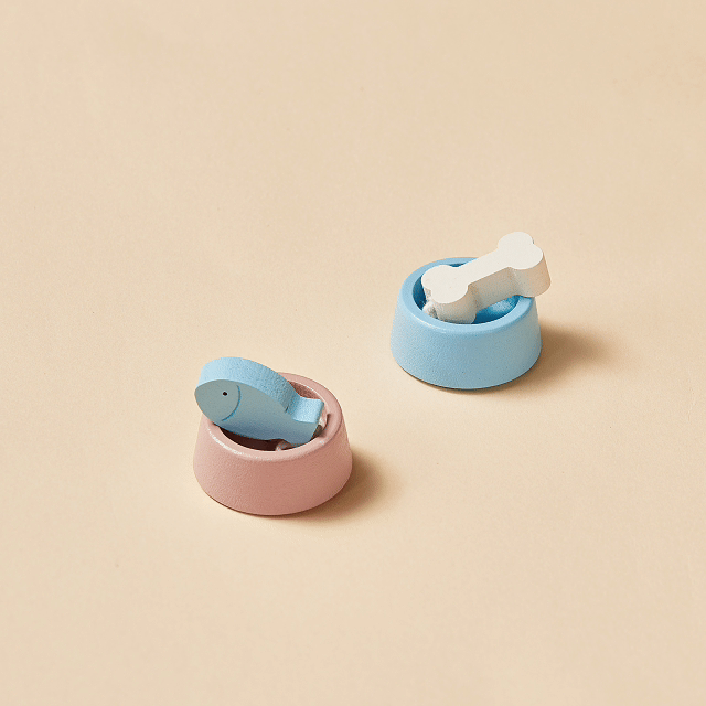 Two small pet bowls from Test's Early Bird Eggs-Tiny Land® Happy Urban Paws playset sit on a beige surface: one pink with a blue fish, one blue with a white bone—perfect pretend pet toys for any mini pet house.