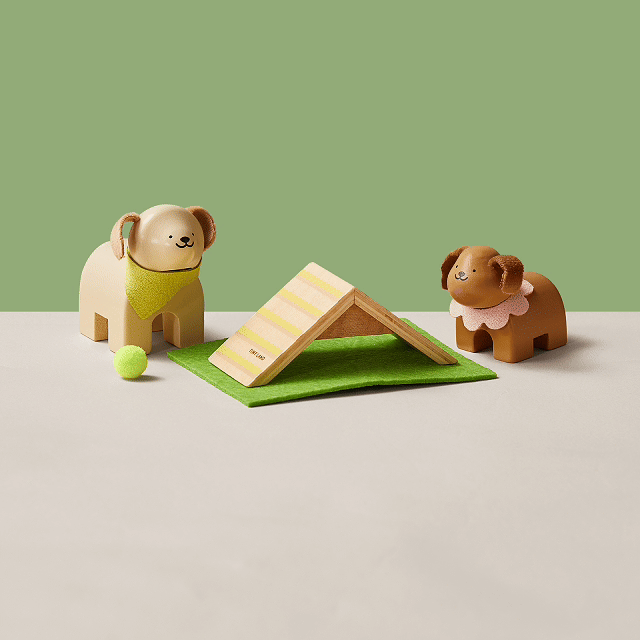 Two Early Bird Eggs-Tiny Land® Happy Urban Paws wooden dog figurines by Test face each other on a beige surface, with a green mat, yellow-striped obstacle, and small tennis ball between them. The background is light green.