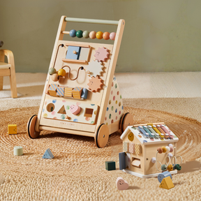 The Tiny Land Complete Development Playset — Baby Walker + Activity Cube is shown on a woven rug, with its activity features and sensory toy house featuring a xylophone and colorful shapes, plus a small wooden chair in the background.