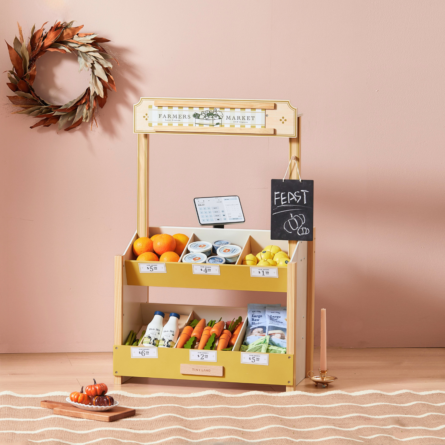 The Tiny Land INC Farmers’ Market Gift Set features a market stall, organic food toys, price tags, a "FEAST" chalkboard sign, and autumn decor—perfect for pretend play on a light wood floor with a pink wall backdrop.