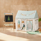 A cozy children’s playroom with the Tiny Land Nature-inspired Play & Art Set—Sage Easel + Forest Play House—plus a small basket and soft toys arranged on a wooden floor.