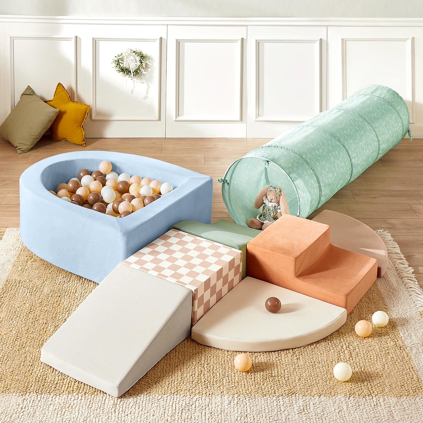 The Tiny Land® PlayNest Climbing Ball Pit Set with Tunnel includes a blue ball pit with neutral balls, pastel climbing blocks, steps, a ramp, round mats, a green tunnel, and sits on a beige rug. Decorative pillows and wall complete the look.
