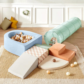 The Tiny Land® PlayNest Climbing Ball Pit Set with Tunnel includes a blue ball pit with neutral balls, pastel climbing blocks, steps, a ramp, round mats, a green tunnel, and sits on a beige rug. Decorative pillows and wall complete the look.