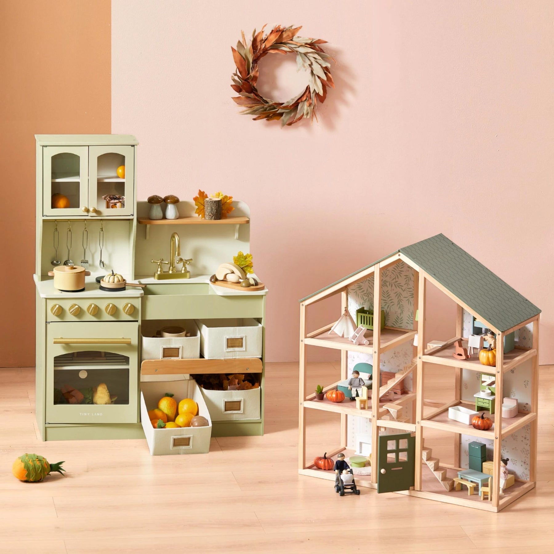The Tiny Land Little Green World Gift Set, a sage green play kitchen for kids with shelves, drawers, pretend food, and utensils, stands by a wooden dollhouse. An autumn wreath decorates the pink wall—perfect for imaginative play.