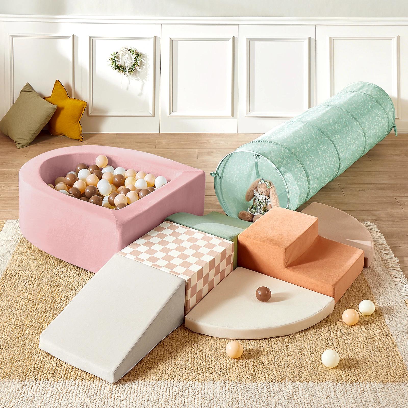 The Tiny Land® PlayNest Climbing Ball Pit Set with Tunnel by Tiny Land features a pastel indoor playground, including a pink foam ball pit with multicolored balls, climbing blocks, green tunnel, orange steps, and cushions on a beige rug.