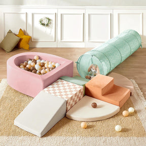 The Tiny Land® PlayNest Climbing Ball Pit Set with Tunnel by Tiny Land features a pastel indoor playground, including a pink foam ball pit with multicolored balls, climbing blocks, green tunnel, orange steps, and cushions on a beige rug.