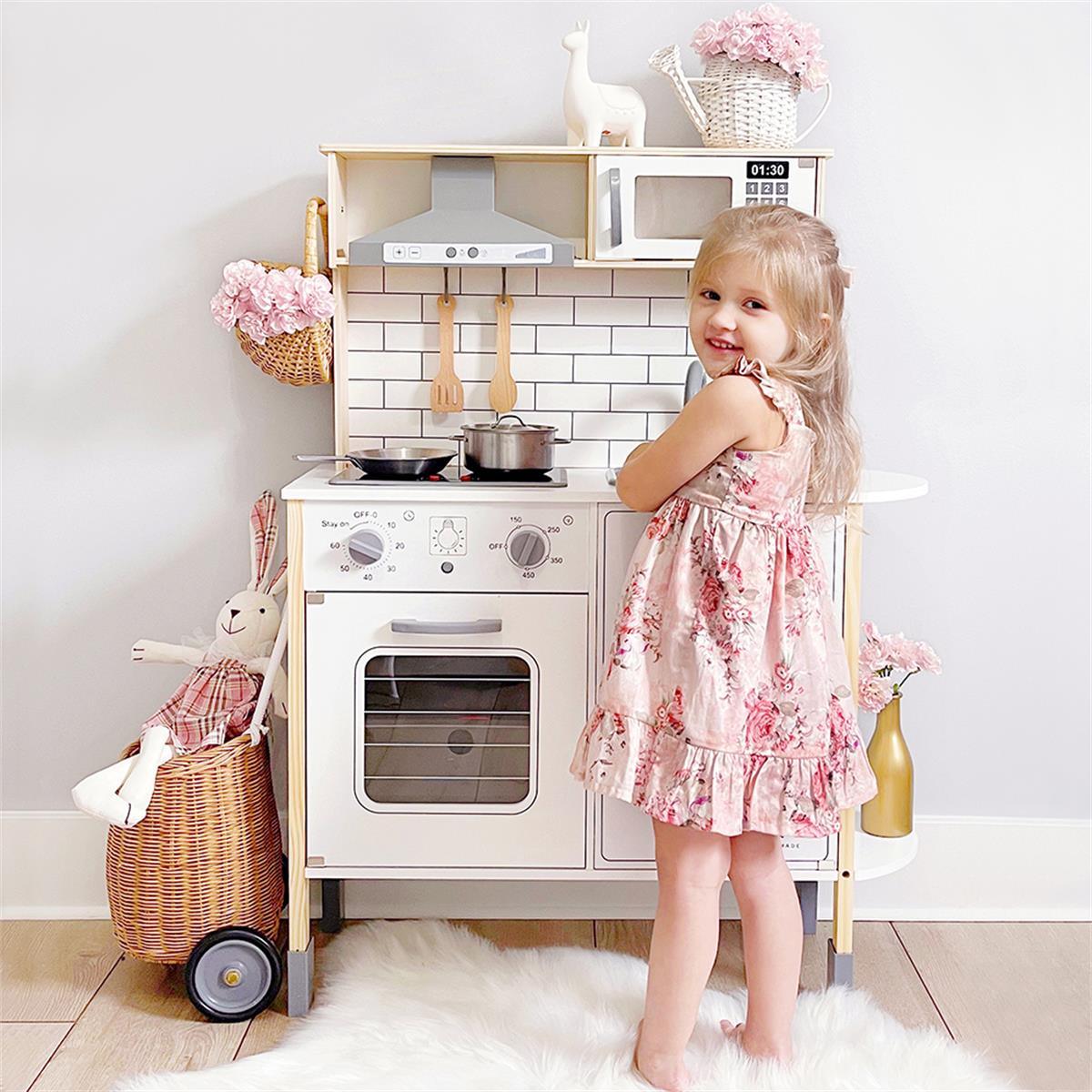 A young girl smiles while playing at the Tiny Land® Farm-to-Table Gift Set — Light & Sound Kitchen & Market Stall. A plush bunny sits in a basket nearby, and the play kitchen features pink flowers and white accessories.
