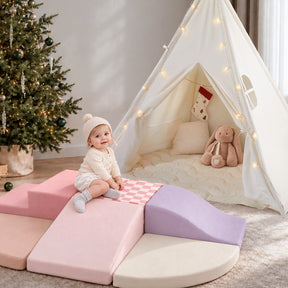 A toddler sits on soft, modular Violet PlayNest Climbing Blocks in a play corner in front of a white Tiny Land® Teepee Tent decorated with fairy lights.