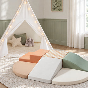 A white fabric Tiny Land® Teepee Tent & PlayNest Climbing Blocks set with string lights next to soft modular climbing blocks in cream, peach, and sage green, creating a cozy play corner.