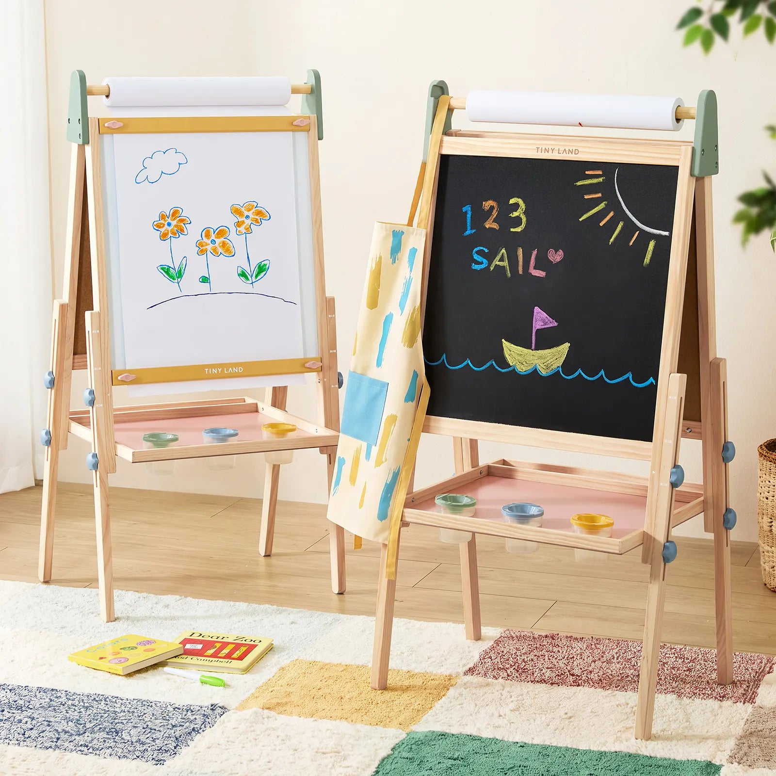 Two Tiny Land® 3-in-1 Art Easels for Kids by Tiny Land stand side by side; one displays a drawing of flowers, while the other features “123 SAIL” and a boat on the chalkboard. Art supplies are organized on shelves beneath the adjustable easel, with a colorful rug and books placed on the floor nearby.|green